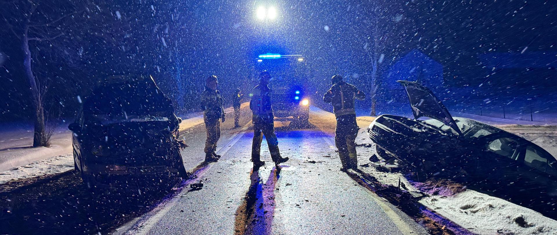 Zdjęcie wykonane nocą pod światło reflektorów na miejscu wypadku w trudnych warunkach zimowych. Intensywnie sypiący śnieg jest rozświetlony przez niebieskie sygnały błyskowe wozu strażackiego stojącego w oddali. Na pierwszym planie, po obu stronach drogi, widać sylwetki dwóch rozbitych samochodów oraz trzech strażaków stojących na jezdni. Silne światło z tyłu tworzy efekt kontrastu, podkreślając padający śnieg i zniszczenia pojazdów. Cała okolica jest spowita mrokiem i bielą śniegu.