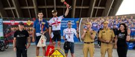 Podium kolarskiego wyścigu z trzema zwycięzcami – na najwyższym stopniu stoi mężczyzna w czarnej czapce i koszulce, trzymający trofeum nad głową, obok niego na drugim i trzecim miejscu stoją kolejni zawodnicy z medalami na szyjach. W tle baner zawodów kolarskich oraz grupa osób, w tym dwóch mężczyzn w mundurach państwowej straży pożarnej i kobieta w czarnej sukience z logo rowerowym. Na podium dekoracja z rowerowymi kołami.