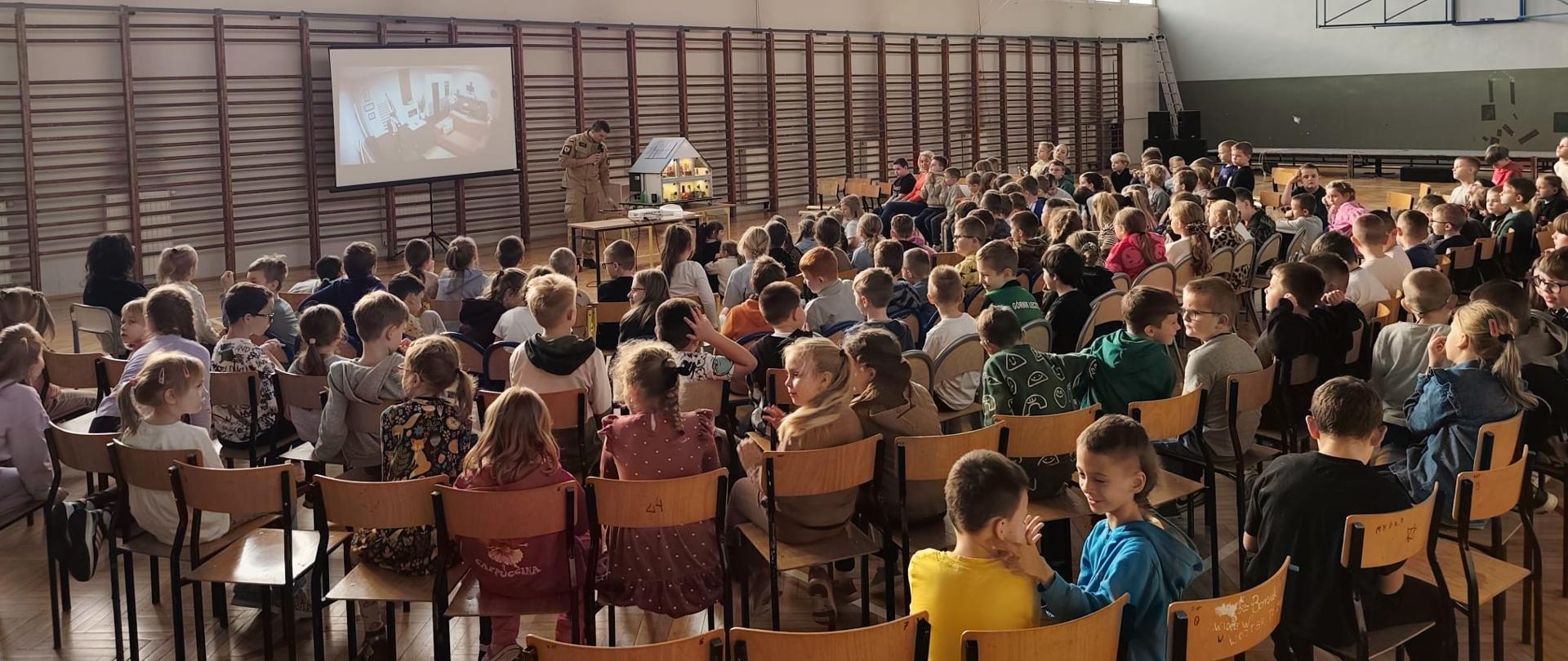 Na zdjęciu widać salę gimnastyczną wypełnioną dziećmi siedzącymi na krzesłach, ustawionych w rzędach. Przed dziećmi stoi funkcjonariusz w mundurze PSP obok którego na stoliku stoi makieta domu. Za nim znajduje się ekran z rzutnikiem na którym wyświetlony jest obraz. Sala jest jasna z dużymi oknami. Dzieci wyglądają na skupione. 