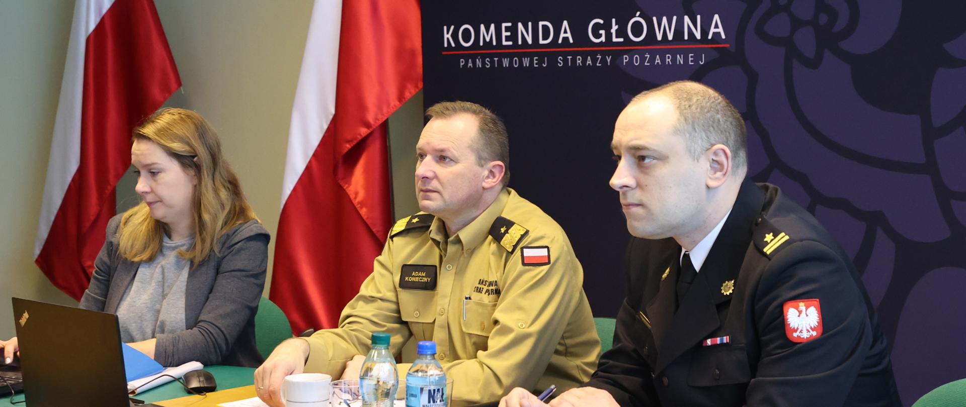 Dwaj strażacy spoglądający na ustawiony przed nimi ekran oraz jedna pani w ubraniu cywilnym w trakcie wideokonferencji. 