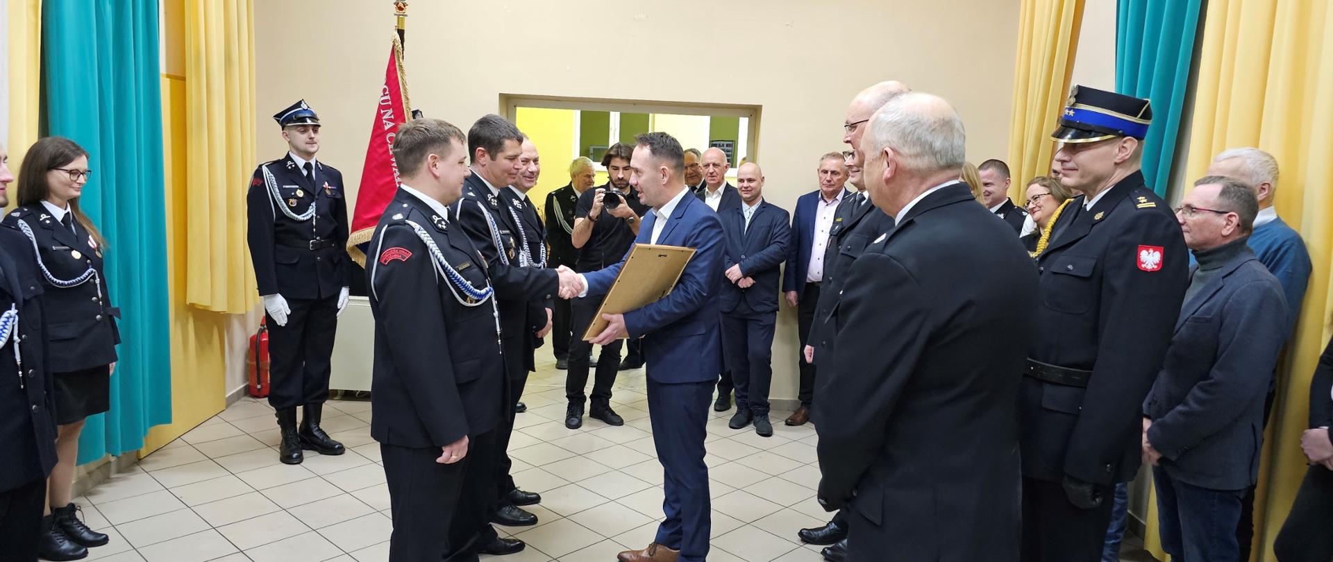Zbiórka strażaków w galowych mundurach w jasnym pomieszczeniu. Wręczenie aktu i wymiana uścisku dłoni mężczyzny w garniturze ze strażakiem stojącym szeregu.