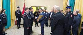 Zbiórka strażaków w galowych mundurach w jasnym pomieszczeniu. Wręczenie aktu i wymiana uścisku dłoni mężczyzny w garniturze ze strażakiem stojącym szeregu.