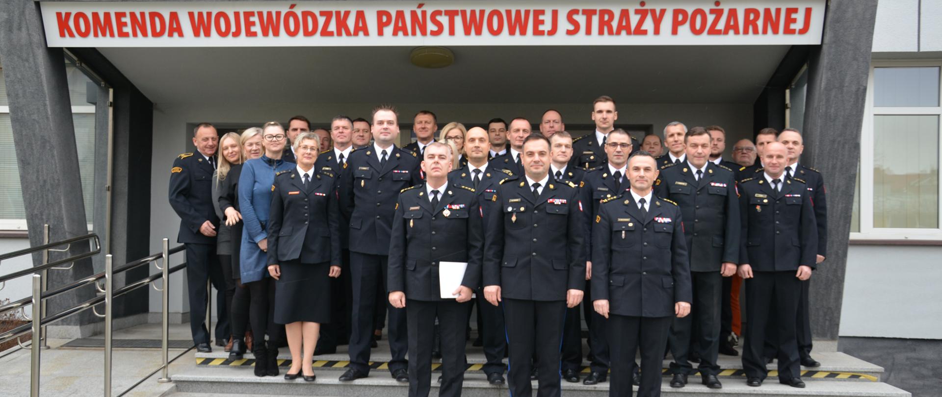 Na zdjęciu widzimy grupę funkcjonariuszy w mundurach oraz pracowników cywilnych stojących na schodach przed wejściem do KW PSP Kielce. Na ich głowami napis "Komenda Wojewódzka Państwowej Staży Pożarnej".