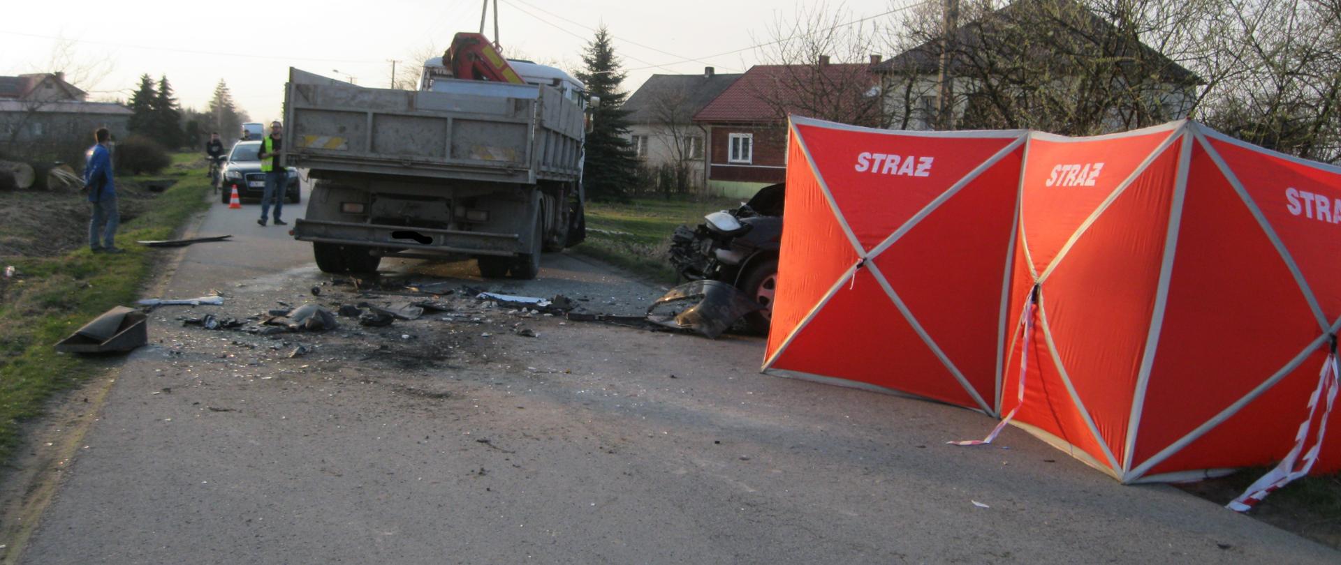 na miejscu widoczny samochód osobowy ,który leży w rowie z widocznymi uszkodzeniami karoserii częściowo osłonięty parawanem oraz uszkodzony samochód ciężarowy ,który blokuje pas jezdni