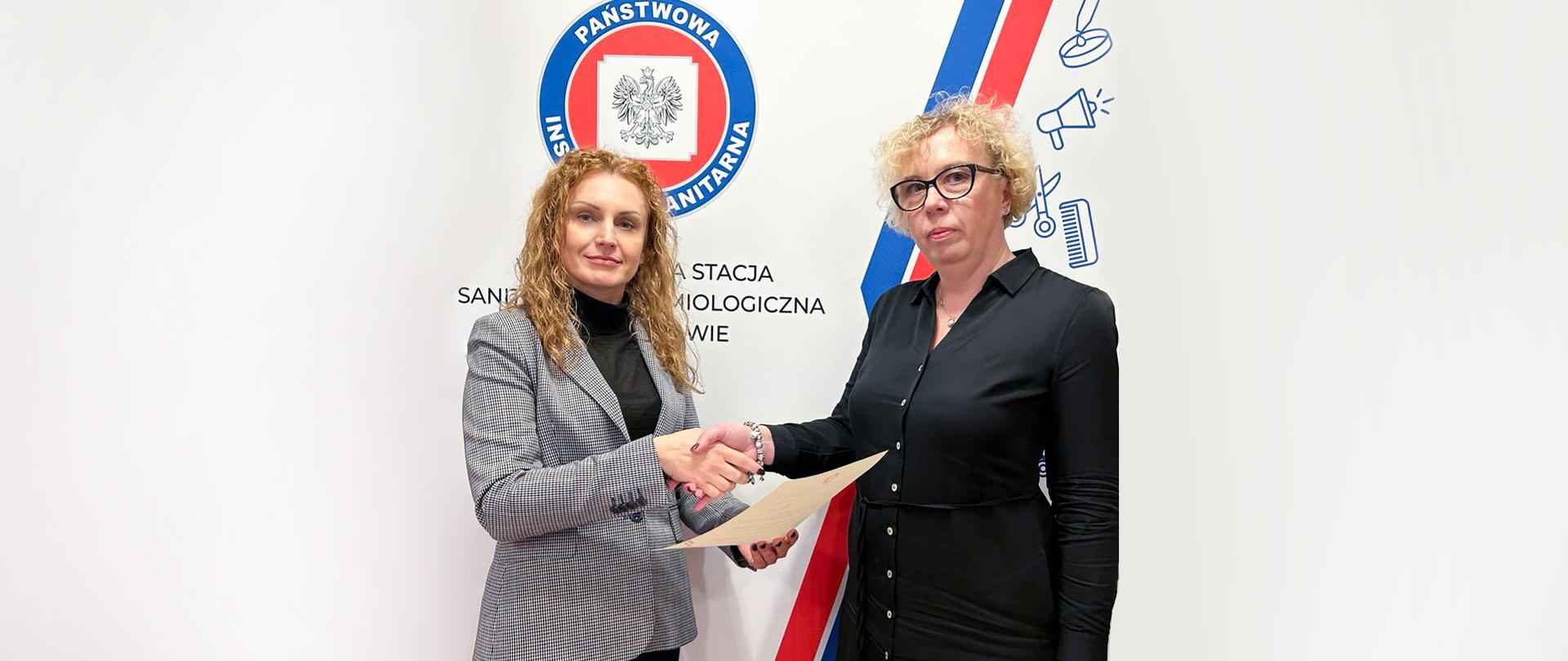 Dwie kobiety ściskają sobie ręce. Kobieta w żakiecie wręcza pismo z powołaniem kobiecie w sukience. W tle logo Inspekcji Sanitarnej.