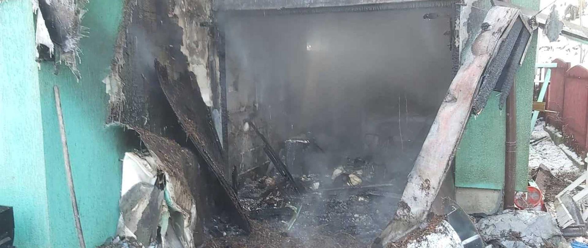 Na zdjęciu widoczna jest opalona, okopcona fasada garażu wbudowanego w budynek mieszkalny. Widoczna jest zniszczona przez pożar zielona elewacja budynku oraz wypalona framuga drzwi garażowych. W głębi zdjęcia widać doszczętnie spalone wnętrze garażu wraz z jego wyposażeniem. Pod ścianami budynku widać różnego rodzaju elementy wyposażenia takie jak wiadra, grabie oraz zniszczone elementy budynku takie jak pokrycie dachu. Z wnętrza garażu widoczny jest wydobywający się dym częściowo zasłaniający widok garażu. 