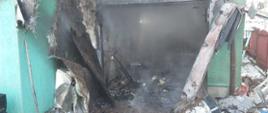 Na zdjęciu widoczna jest opalona, okopcona fasada garażu wbudowanego w budynek mieszkalny. Widoczna jest zniszczona przez pożar zielona elewacja budynku oraz wypalona framuga drzwi garażowych. W głębi zdjęcia widać doszczętnie spalone wnętrze garażu wraz z jego wyposażeniem. Pod ścianami budynku widać różnego rodzaju elementy wyposażenia takie jak wiadra, grabie oraz zniszczone elementy budynku takie jak pokrycie dachu. Z wnętrza garażu widoczny jest wydobywający się dym częściowo zasłaniający widok garażu. 