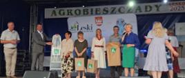 Na tle plakatu znajduje się grupa osób, od lewej Sekretarz Powiatu leskiego Czesław Gawłowski, Starosta Powiatu Leskiego Wojciech Stelach, nagrodzeni w konkursie, którzy w rękach trzymają otrzymane nagrody oraz kierownik PT KRUS Sanok Gabriela Bałut.