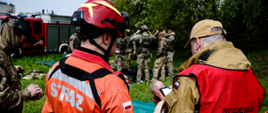 Strażak z grupy poszukiwawczej stoi obok strażaka rozjemcy niedaleko stoją policjanci.