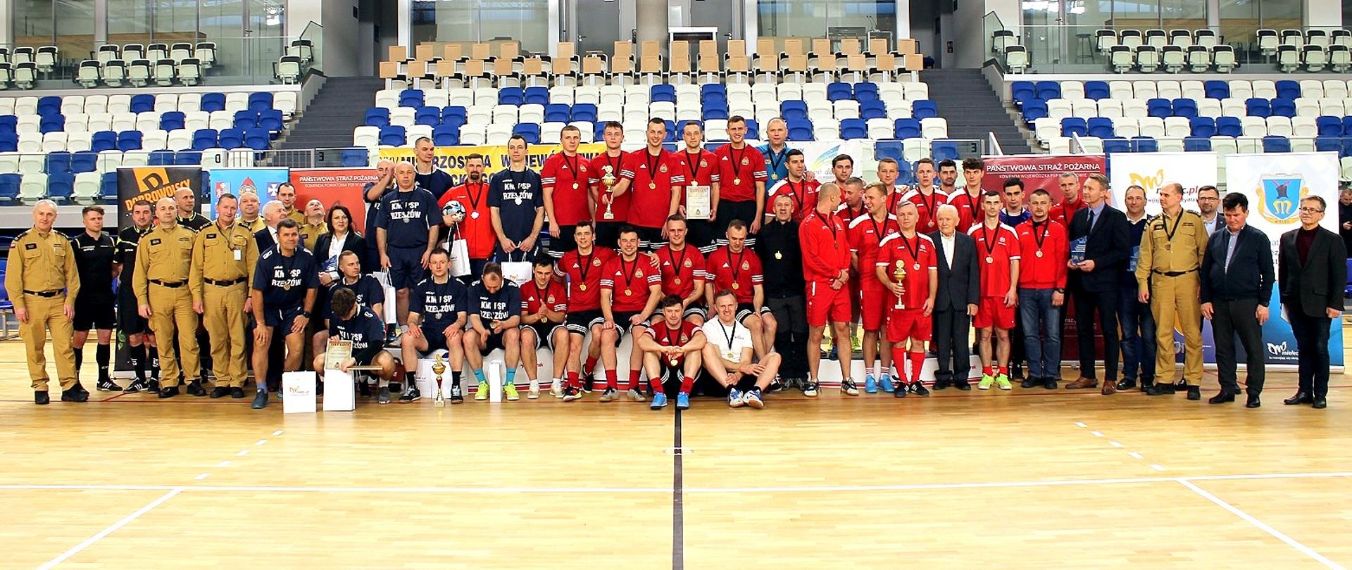 Fotografia wykonana podczas zawodów finałowych XXV Mistrzostwa Województwa Podkarpackiego Strażaków PSP w Futsalu. W szeregach stoją funkcjonariusze i goście zaproszeni na finał zawodów. Na środku fotografii stoją na podium zawodnicy. Za osobami widać banery organizatorów i sponsorów.