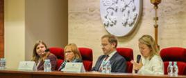 Przy stole prezydialnym siedzą cztery osoby, trzy kobiety i jeden mężczyzna. Na stole stoi woda oraz standy z imionami i nazwiskami prelegentów, a także mikrofon. Kobieta po prawej stronie trzyma mikrofon oraz przemawia do uczestników.