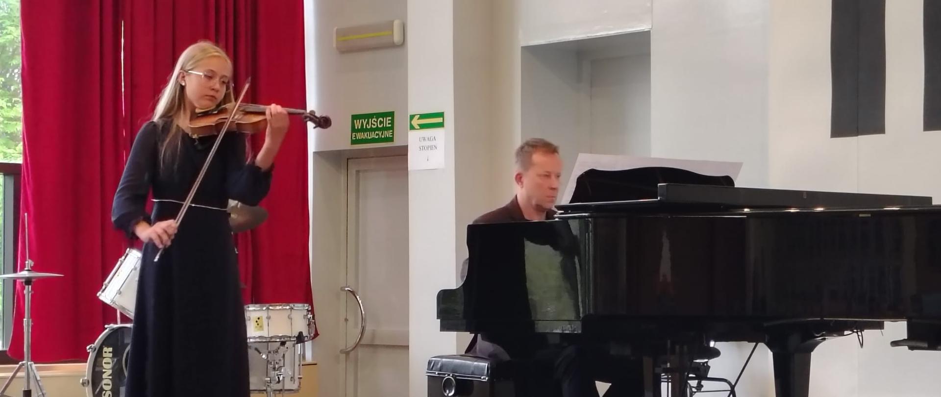Dziewczynka gra na skrzypcach, mężczyzna akompaniuje jej na fortepianie.