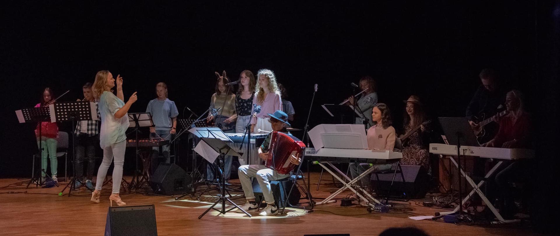 Zespół uczniów gra na różnych instrumentach takich jak akordeon, keyboard, wibrafon, flet, gitara. Przed kobieta, która im dyryguje.