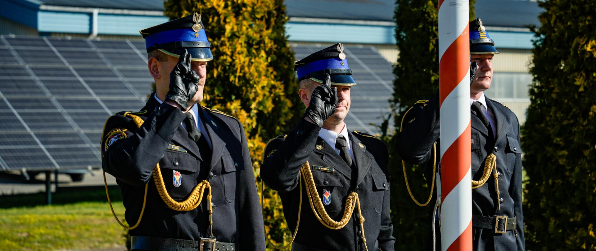 Podniesienie flagi Rzeczypospolitej Polskiej podczas uroczystej zbiórki z okazji Dnia Flagi w Komendzie Powiatowej PSP w Lublińcu; funkcjonariusze w galowych mundurach oddają honory, na tle budynku i paneli fotowoltaicznych.