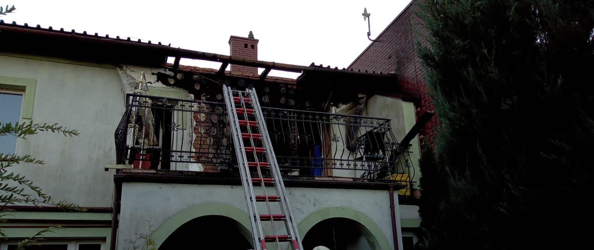 Zdjęcie przedstawia budynek, w którym na pierwszej kondygnacji doszło do pożaru. Widoczna spalona elewacja oraz uszkodzenie dachu. Poniżej oparta o balustradę balkonu znajduje się drabina wysuwana oraz trzech strażaków.