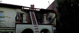 Zdjęcie przedstawia budynek, w którym na pierwszej kondygnacji doszło do pożaru. Widoczna spalona elewacja oraz uszkodzenie dachu. Poniżej oparta o balustradę balkonu znajduje się drabina wysuwana.