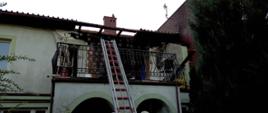 Zdjęcie przedstawia budynek, w którym na pierwszej kondygnacji doszło do pożaru. Widoczna spalona elewacja oraz uszkodzenie dachu. Poniżej oparta o balustradę balkonu znajduje się drabina wysuwana.
