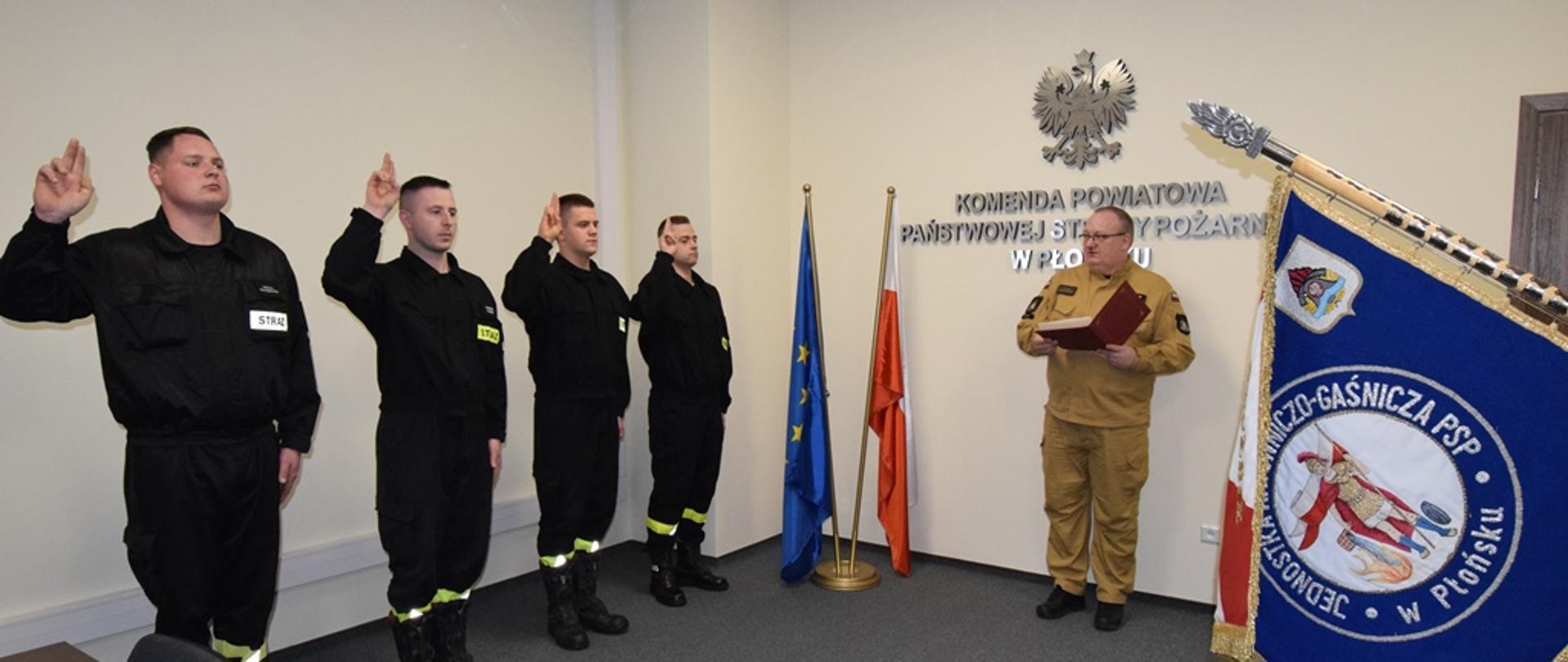 Z lewej strony czterech strażaków ubranych w czarne ubrania koszarowe, z podniesioną prawą ręką ślubują. Na środku oficer Państwowej Straży Pożarnej w umundurowaniu służbowym w kolorze piaskowym odczytuje rotę ślubowania. Z prawej strony poczet sztandarowy. 