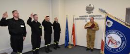 Z lewej strony czterech strażaków ubranych w czarne ubrania koszarowe, z podniesioną prawą ręką ślubują. Na środku oficer Państwowej Straży Pożarnej w umundurowaniu służbowym w kolorze piaskowym odczytuje rotę ślubowania. Z prawej strony poczet sztandarowy. 