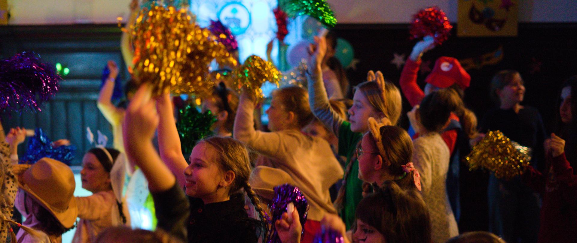 Grupa dzieci bawi się i tańczy w kolorowo oświetlonej sali, trzymając błyszczące pompony w różnych kolorach. Uczestnicy mają na sobie różne kostiumy i opaski, a atmosfera przypomina radosną zabawę lub szkolną imprezę.