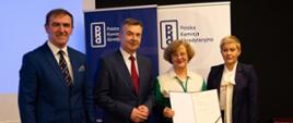Na tle baneru z napisami Polska Komisja Akredytacyjna stoją od lewej mężczyzna w granatowym garniturze, minister Wieczorek, trzymająca otwarty dokument kobieta w biało-zielonej bluzie i wiceminister Mrówczyńska.