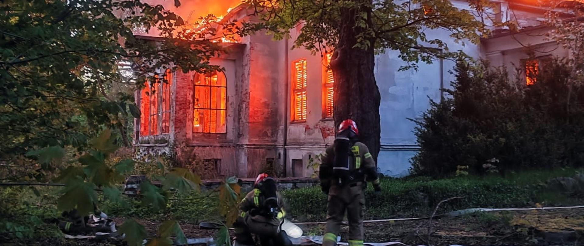 Dwaj strażacy w ubraniu specjalnym rozwijają węże, w tle palący sie budynek szkoły, z okien wydostają się płomienie.