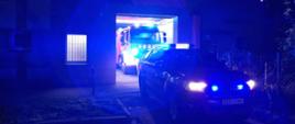 Jeden samochód pożarniczy stoi przed budynkiem za nim drugi - w środku. Oba mają włączone światła błyskowe. Sceneria nocna.