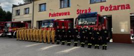 Plac manewrowy przed komenda w Rawiczu. Na tle budynku i stojących przed nią wozów bojowych, w szeregu ustawieni są strażacy. Ci stojący z lewej strony mają na sobie umundurowanie służbowe w kolorze piaskowym, ci z prawej - koszarowe w kolorze czarnym. Na budynku, nad bramami garazowymi widnieje czerwony napis PAŃSTWOWA STRAŻ POŻARNA