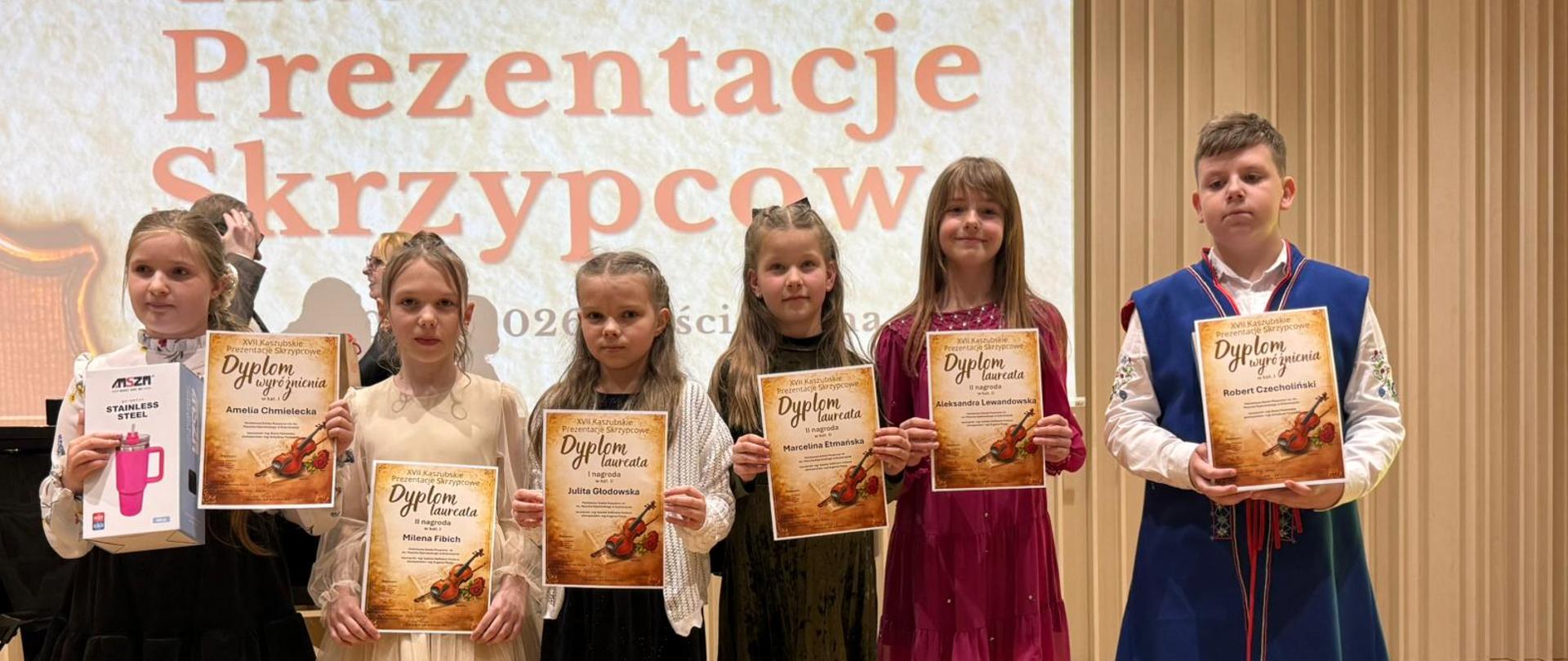 Na zdjęciu sześcioro młodych skrzypków, którzy zostali laureatami konkursu. Wszyscy ubrani odświętnie. Każdy z nich trzyma dyplom. W tle brązowy napis: Kaszubskie Prezentacje Skrzypcowe.