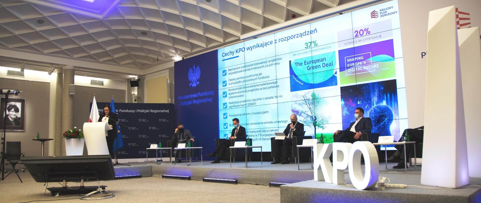 W sali konferencyjne na podeście siedzi w fotelach 5 mężczyzn. Drugi od lewej wiceminister Waldemar Buda. Za nimi ekran z wyświetloną prezentacją. Z lewej strony w mównicy stoi wiceminister Małgorzata Jarosińska-Jedynak. Przed nią na podłodze monitor. Z boku flagi PL i UE.