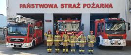 Widok z przodu. Sześciu strażaków JRG Andrychów stoi w ubraniach specjalnych przed samochodami pożarniczymi przed garażami JRG. 