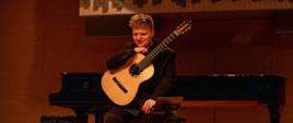 Gitarzysta siedzący na krześle, trzymający gitarę klasyczną - opartą na podgitarniku opartym na prawej nodze, prawą rękę opiera na górnej części korpusu instrumentu, a na niej podpiera brodę. Za plecami widoczny fortepian, a z tyłu na górze prospekt organowy - scena sali koncertowej szkoły