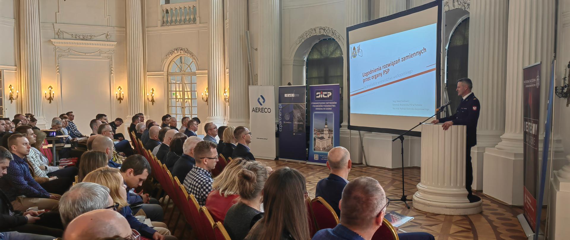 Strażak w mundurze przemawia przez mikrofon stojąc przy mównicy. Obok widoczny ekran z prezentacją oraz osoby siedzące na krzesłach.
