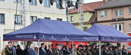 W centrum kadru, przy czerwonej mównicy z logotypem Państwowej Straży Pożarnej, stoi oficer w ciemnym mundurze galowym, który wygłasza mowę. Za nim, pod dużymi granatowymi namiotami z napisami „Państwowa Straż Pożarna”, zgromadzeni są goście honorowi w strojach wizytowych, duchowni oraz liczne rzędy umundurowanych strażaków. W tle widoczne są jasne fasady kamienic oraz profesjonalne konstrukcje oświetleniowe.