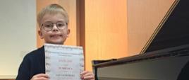 Na zdjęciu chłopiec, blondyn w okularach i granatowym garniturze. Chłopiec siedzi przy czarnym fortepianie i trzyma dyplom. W oddali napis "Malborskie Prezentacje Fortepianowe Pierwszaki na Estradzie". Tło obrazka w kolorze jasno brązowym.