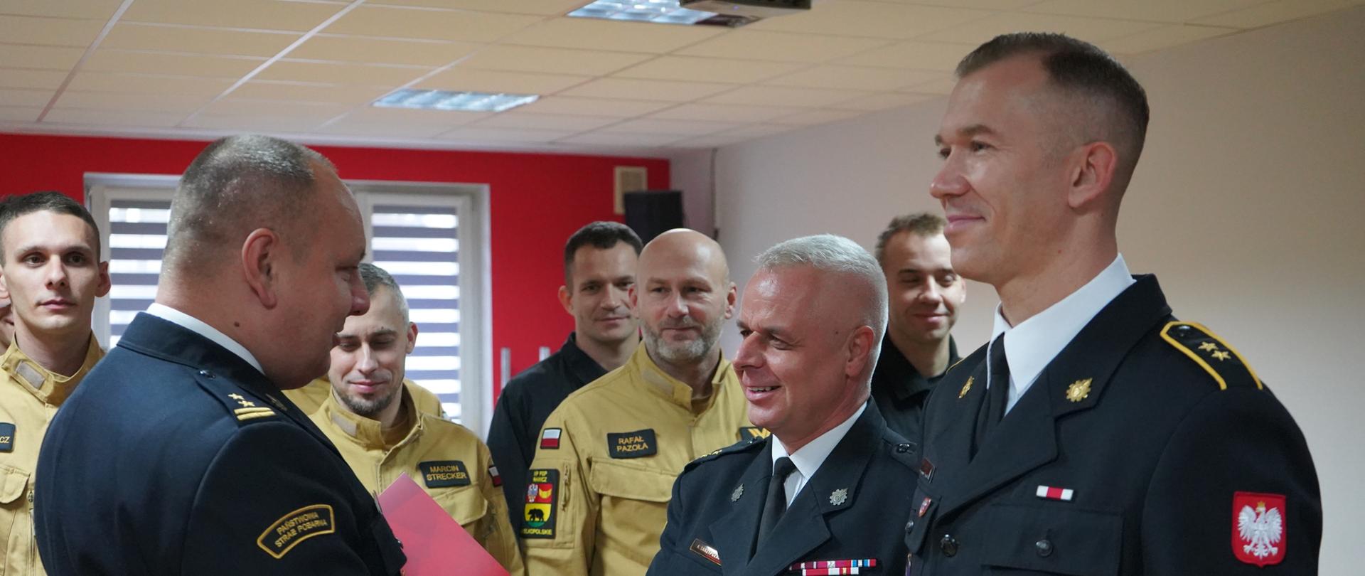 Na zdjęciu widać uroczystość wręczenia awansów i wyróżnień w Komendzie Powiatowej Państwowej Straży Pożarnej w Rawiczu. Strażacy w galowych mundurach odbierają gratulacje i uściski dłoni od przełożonych. W tle widoczny jest emblemat Państwowej Straży Pożarnej oraz napis z nazwą komendy. Zdjęcie oddaje podniosły i uroczysty charakter wydarzenia.