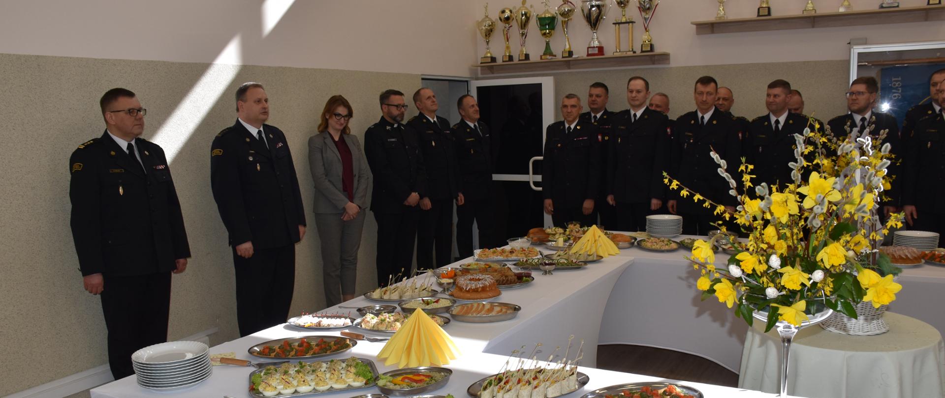 Strażacy i zaproszeni goście stojący wokół stołu wielkanocnego