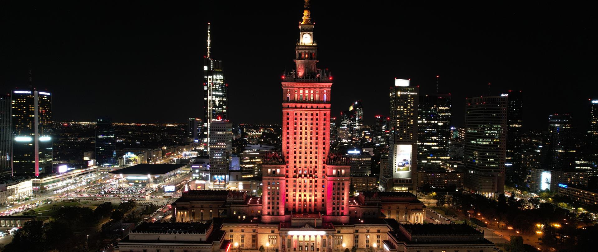 Na zdjęciu wykonanym wieczorną porą widać Pałac Kultury i Nauki podświetlony na pomarańczowo (barwy Światowego Dnia Bezpieczeństwa Pacjenta). W tle rozciąga się panorama wieżowców. 