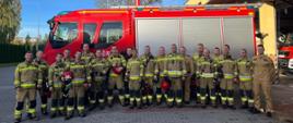 Wspólne zdjęcie strażaków żegnających st. ogn. Marcina Lechowskiego przechodzącego na zaopatrzenie emerytalne na tle samochodu pożarniczego. Strażacy stoją ubrani w ubrania specjalne, 