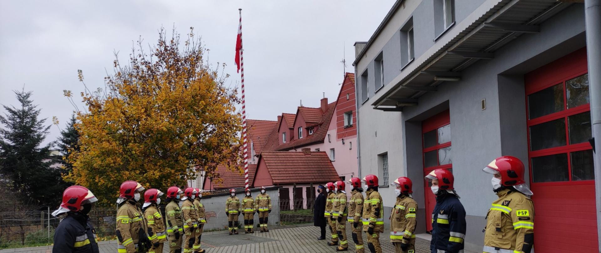 Uroczyste podniesienie flagi państwowej na maszt. Strażacy Komendy Powiatowej Państwowej Straży Pożarnej w Lęborku w ubraniach specjalnych i hełmach strażackich stoją w dwóch rzędach naprzeciw siebie. Na maszcie powiewa flaga państwowa. W tle przy maszcie stoi poczet flagowy składający się z trzech strażaków. Ze strażakami stoi Zastępca Komendanta Powiatowego Państwowej Straży Pożarnej w Lęborku w umundurowaniu. Strażacy stoją na baczność. Z powodu pandemii koronawirusa wszyscy mają założone maseczki ochronne. 