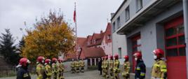 Uroczyste podniesienie flagi państwowej na maszt. Strażacy Komendy Powiatowej Państwowej Straży Pożarnej w Lęborku w ubraniach specjalnych i hełmach strażackich stoją w dwóch rzędach naprzeciw siebie. Na maszcie powiewa flaga państwowa. W tle przy maszcie stoi poczet flagowy składający się z trzech strażaków. Ze strażakami stoi Zastępca Komendanta Powiatowego Państwowej Straży Pożarnej w Lęborku w umundurowaniu. Strażacy stoją na baczność. Z powodu pandemii koronawirusa wszyscy mają założone maseczki ochronne. 