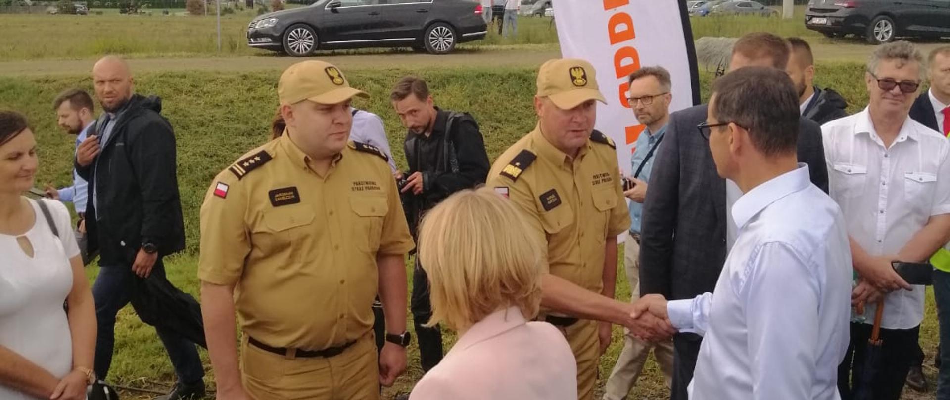 grupa ludzi sto na trawniku, mężczyzna w mundurze strażackim podaje rękę mężczyźnie w białej koszuli i czarnych stpodniach, obok stoi drugi mężczyzna w mundurze strażaka, w tle kilka osób oraz dwa czarne samochody