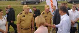 grupa ludzi sto na trawniku, mężczyzna w mundurze strażackim podaje rękę mężczyźnie w białej koszuli i czarnych stpodniach, obok stoi drugi mężczyzna w mundurze strażaka, w tle kilka osób oraz dwa czarne samochody