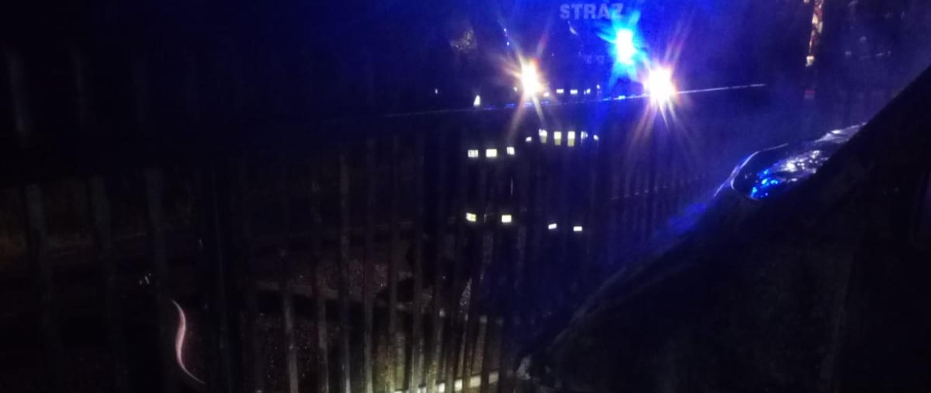 Na zdjęciu wykonanym w porze nocnej widoczny jest przód autobusu, który uległ spaleniu oraz brama. Za ogrodzeniem stoi samochód strażacki oświetlony niebieskimi lampami sygnalizacyjnymi.