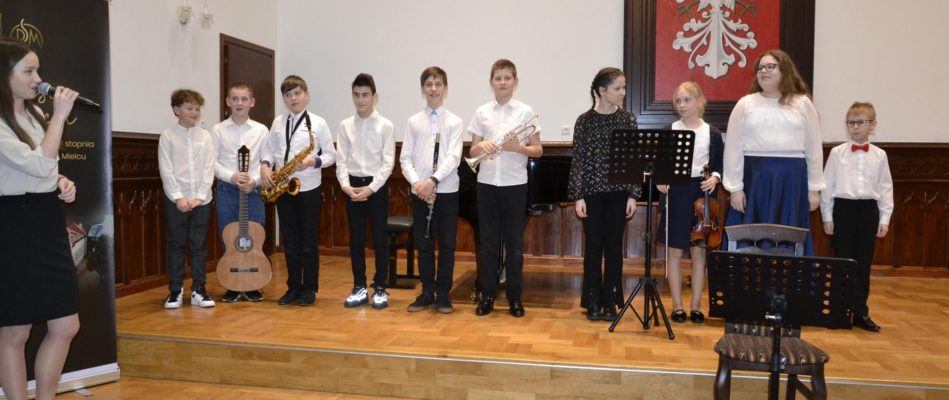 zdjęcie przedstawia uczniów klasy II cyklu czteroletniego PSM I stopnia stojących na scenie Sali Królewskiej w wielkim godłem polski w tle, z lewej strony stoi prowadząca koncert Kalina Rokosz
