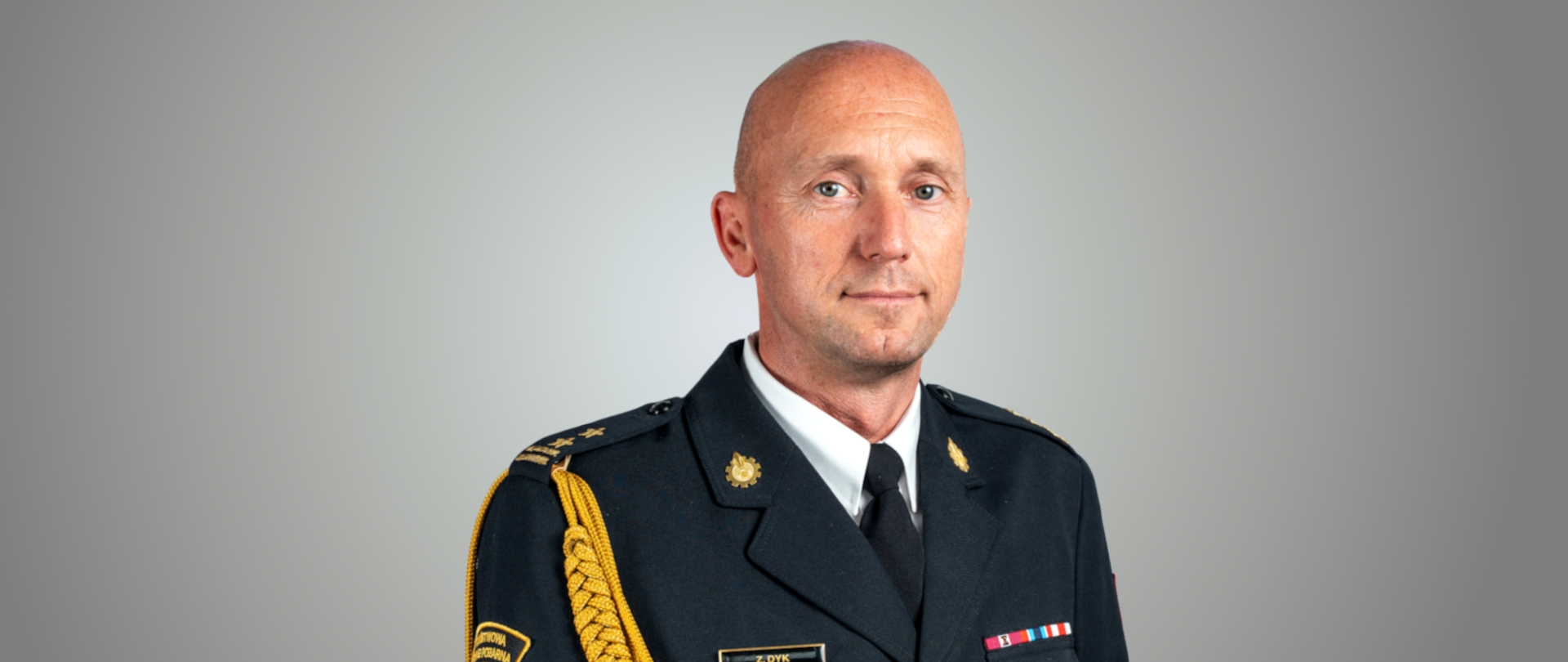 Na zdjęciu znajduje się portret mężczyzny - bryg. Zbigniewa Dyka. Mężczyzna ma owalną twarz i niebieskie oczy. Ubrany jest w ciemny mundur wyjściowy z odznaczeniami, stopniami i naszywką na ramieniu z napisem „PAŃSTWOWA STRAŻ POŻARNA”.
