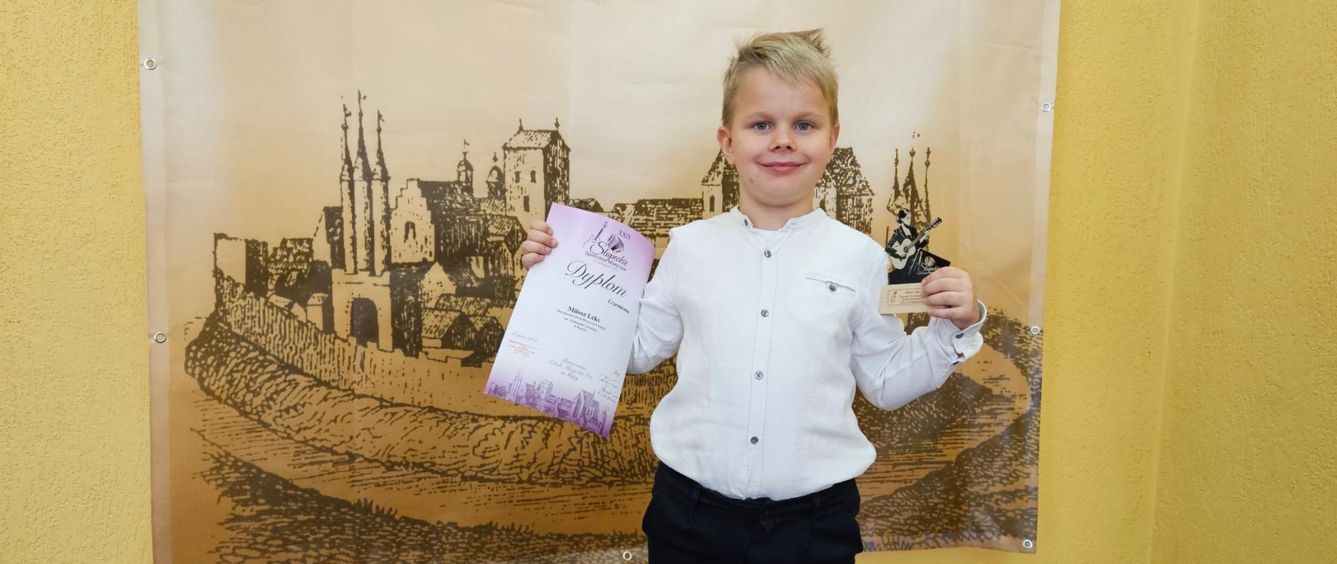 Na zdjęciu uczeń w stroju galowym pozujący z dyplomem i statuetką na tle banneru Słupeckich Spotkań Muzycznych. 