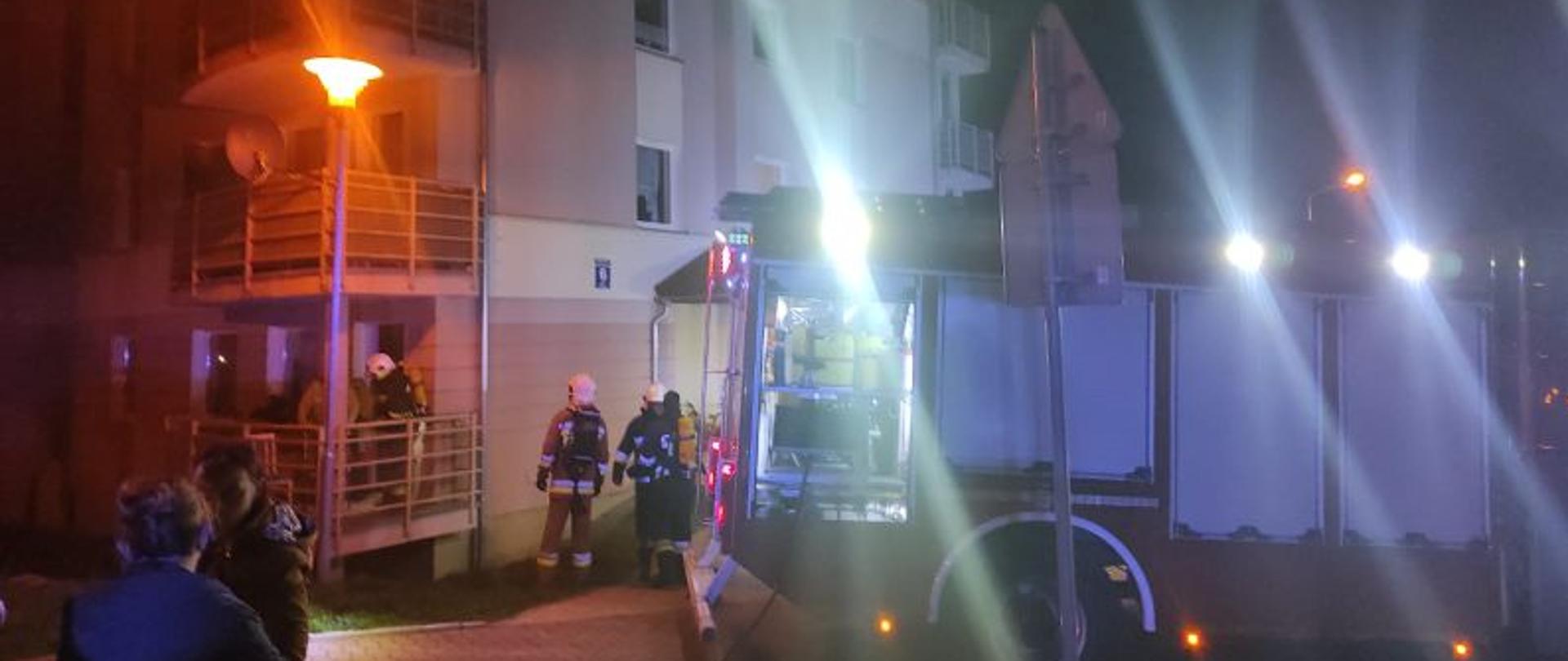 Działania straży pożarnej po zmierzchu. Jest ciemno. Miejsce zdarzenia rozświetlają białe lampy pojazdu gaśniczego. Pojazd stoi przy budynku bokiem do balkonu. Balkon znajduje się na parterze. Na balkonie stoją strażacy i dogaszają spalone meble