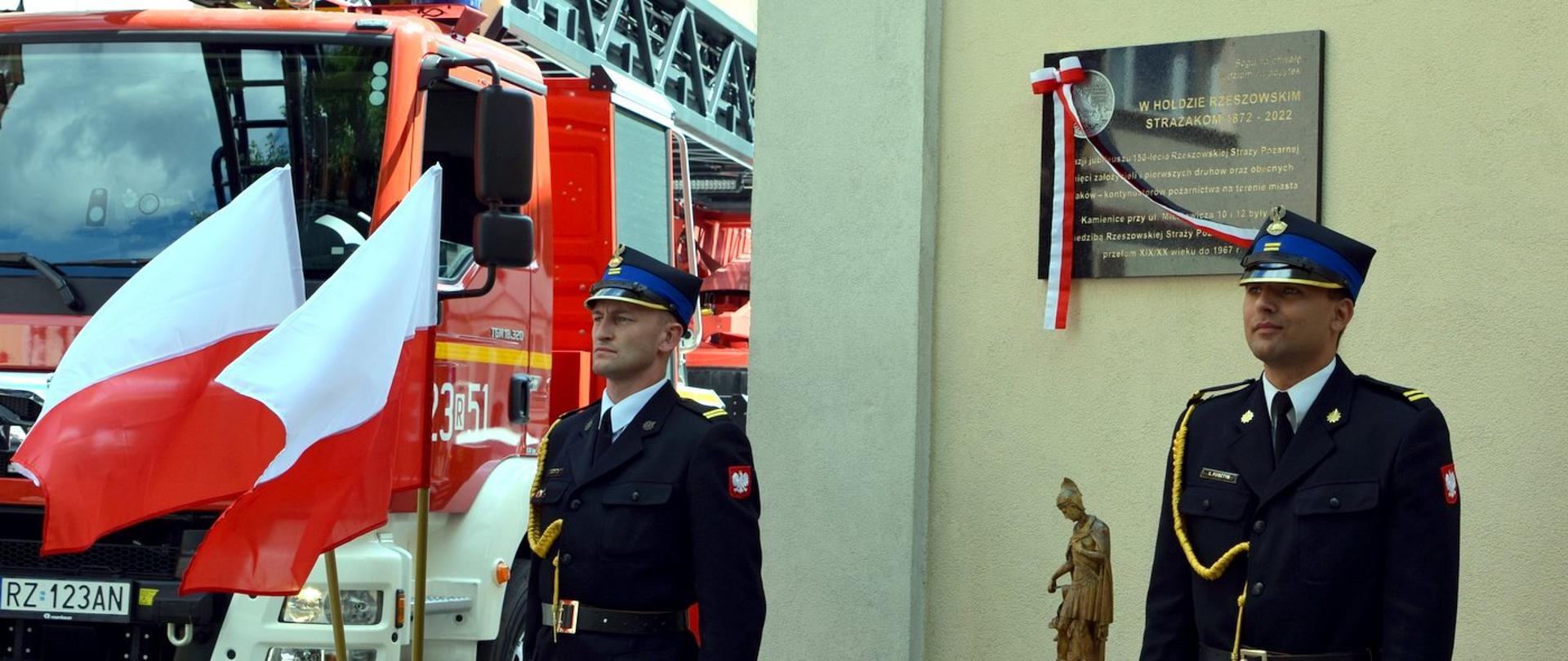 Dwóch strażaków Państwowej Straży Pożarnej pełni Wartę honorową przy tablicy pamiątkowej. W tle widać samochód pożarniczy.