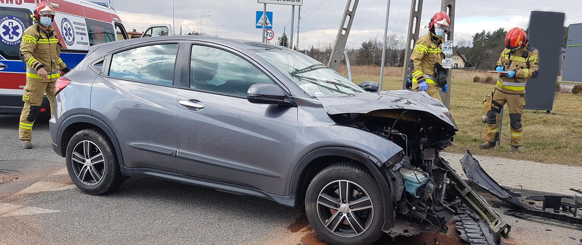 Prawy bok szarego samochodu osobowego z rozbitym przodem. Za samochodem trzech strażaków oraz karetka pogotowia. Z samochodu na drogę wylał się płyn.